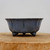 A high-resolution, side-view studio photograph of a glazed Yixing bonsai pot, model YX905, resting on a thick, light-grained wooden slab. The pot is finished in a sophisticated, dusty blue-grey glaze with a subtle, fine-grained texture. Its deep, round body features elegant vertical indentations that create a lobed, petal-like effect around its circumference, leading up to a thin, slightly flared rim. The vessel is elevated on three small, ornate "cloud" feet carved from dark reddish-brown clay, which provides a beautiful contrast to the blue glaze. The minimalist, off-white background draws focus to the pot's refined craftsmanship and classic Japanese-inspired aesthetic.