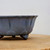 A side-profile close-up of a Yixing YX905 glazed ceramic bonsai pot. The pot features a distinctive lobed or scalloped body with a speckled, lavender-blue glaze that shows subtle variations in color and texture. It has a flared rim and is elevated by three ornate, cloud-patterned tripod feet. The pot is positioned on a smooth, light-grained wooden surface against a minimalist off-white background, highlighting its elegant form and fine craftsmanship.