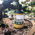 An 8 oz bottle of Uncle Bill's No Nitrogen 0-10-10 liquid fertilizer sits nestled in the coarse, pebbly soil of a mature bonsai tree. The dark plastic bottle, featuring a cartoon farmer on its yellow and green label, is positioned under the thick, textured branch of a bonsai tree with small, vibrant green leaves. 

The label clearly displays the "0-10-10" NPK ratio and states that it "provides a balanced delivery of phosphate (P205) and soluble Potash (K20) to keep your tree's root system healthy and happy." In the background, other potted plants and bright sunlight suggest a greenhouse or nursery setting, emphasizing the product's use in professional bonsai maintenance and care.