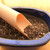 A close-up photograph featuring a light peach-colored plastic soil scoop from a 3-piece set, angled into a deep blue, glossy ceramic bonsai pot. The scoop is filled with a textured bonsai potting mix composed of dark brown bark bits and light-colored grit. The pot itself is partially filled with the same coarse soil blend. This practical demonstration shows the utility of plastic soil scoops for precise potting and repotting of bonsai trees. The scene is set on a light-colored wooden tabletop, emphasizing a clean and organized bonsai gardening workspace.