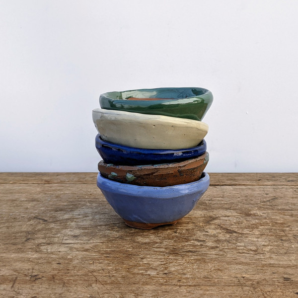 A vertical stack of five small, hand-crafted glazed ceramic pots sits in the center of a rustic, weathered wooden tabletop. The pots, ideal for miniature bonsai trees or succulents, feature unique, slightly irregular shapes and varied finishes. 

From top to bottom, the colors include:
*   A glossy, forest green pot with a wavy rim.
*   A matte, off-white or cream-colored pot.
*   A deep, shiny cobalt blue pot.
*   An earthy, textured terracotta brown pot with subtle green glaze drips.
*   A soft, matte periwinkle blue pot at the base.

The background is a clean, solid off-white wall, which emphasizes the artisanal textures and vibrant colors of the ceramic stoneware. Each pot has a shallow design typical of succulent or bonsai containers.