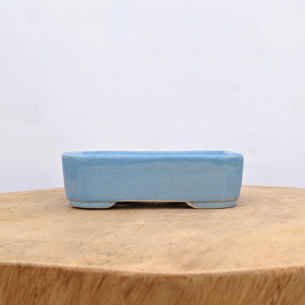 A side-view shot of a five-inch rectangular light blue glazed ceramic bonsai pot. The pot features a subtle, speckled sky-blue glaze with a glossy finish and rounded corners. It sits elevated on two wide, low-profile feet, creating a slight arch underneath for drainage and airflow. The bonsai container rests on a natural, light-colored wooden surface with visible grain patterns, set against a clean, neutral off-white background.