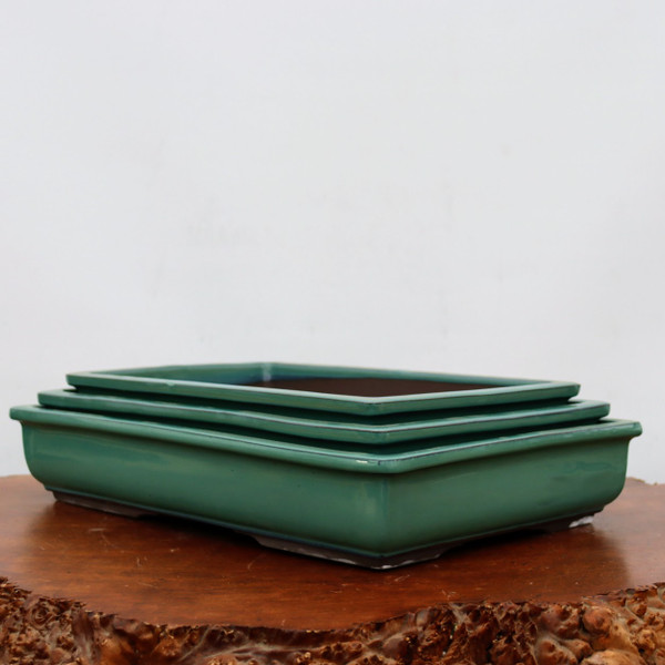 A set of three nesting glazed green bonsai pots, ranging in size and stacked neatly one inside the other. These ceramic planters feature a vibrant, glossy teal-green finish with smooth, rounded corners and a slightly flared rim. The dark brown unglazed interior of the top pot is visible, suggesting a functional design for plant health. The pots are displayed on a polished, dark wood surface with a rough, natural burl edge, all set against a minimalist off-white background. This arrangement highlights the elegant rectangular design and uniform color of these high-quality bonsai containers, ideal for showcasing various tree styles.