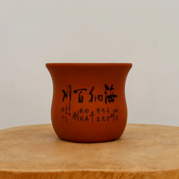 A traditional unglazed reddish-brown terracotta bonsai pot is showcased against a plain, off-white background. The pot features a classic, elegant design with a slightly flared rim and a smooth, rounded body that tapers gently towards the base. It is prominently decorated with intricate black etched Chinese calligraphy across its front. The central, larger characters read "海纳百川" (Hǎi nà bǎi chuān), a profound idiom meaning "The sea can hold the water from a hundred rivers," symbolizing great capacity, tolerance, and inclusivity. Smaller, more delicate lines of poetic script and a traditional artist's seal are etched to the left and below the main phrase. The pot sits on a rustic, light-colored wooden platform, which highlights the earthy, natural texture of the unglazed clay. Soft, even lighting creates subtle highlights on the pot's surface, emphasizing its simple yet sophisticated aesthetic.