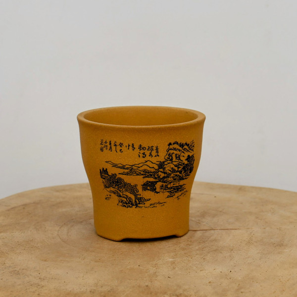A high-angle, eye-level shot features a small, unglazed bonsai pot with a warm, yellow-ochre finish. The ceramic pot has a cylindrical body that tapers slightly toward the base, featuring a subtly flared rim and four small, integrated feet. Centered on its front is a delicate black-ink etched landscape, depicting a tranquil scene of rolling mountains, clusters of pine trees, and a small traditional pavilion nestled near a body of water. To the left of this scene, several vertical columns of elegant Chinese calligraphy are finely inscribed. 

The pot's matte, earthy texture and rich golden-brown color are highlighted by soft, even lighting that casts minimal shadows. It sits on a rustic, light-toned wooden surface with visible grain and natural cracks, adding to the organic feel of the composition. The entire setup is placed against a clean, neutral off-white background, creating a minimalist aesthetic that focuses attention on the detailed craftsmanship of the etched artwork.