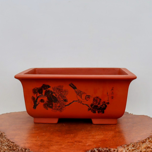 A close-up, eye-level shot of a rectangular unglazed bonsai pot crafted from reddish-brown terracotta clay. The front of the pot is adorned with an intricate, hand-etched scene in black, depicting a small bird delicately perched on a flowering branch. The artwork, which resembles a traditional Chinese ink wash painting, shows detailed peony-like blossoms and leaves with calligraphic markings to the right. The pot has a slightly flared top rim and sits on four sturdy, block-style feet. It is displayed on a polished, warm-toned natural wood slice with live, textured bark edges, all set against a clean, neutral white background. This decorative bonsai container showcases a blend of rustic charm and artistic craftsmanship.