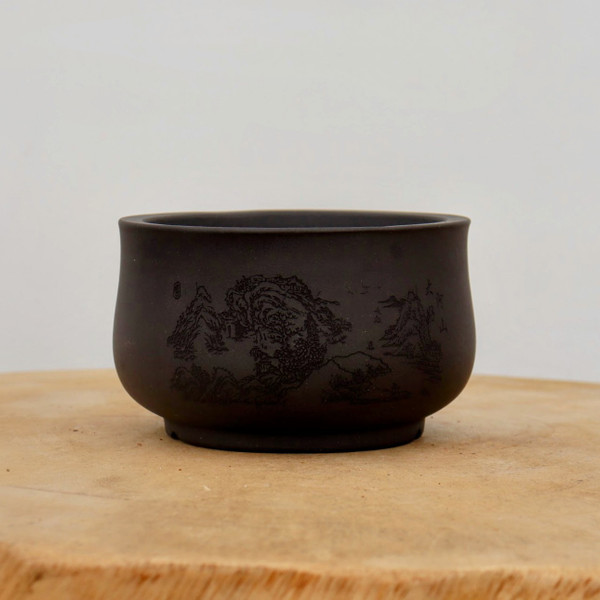 A small, round unglazed ceramic bonsai pot with a dark charcoal-colored matte finish. The front of the planter is adorned with an intricate, finely etched traditional landscape scene featuring high mountain peaks, textured trees, and small hillside buildings. Delicate calligraphic characters are engraved alongside the artwork. The pot has a slightly flared rim and sits on a small, recessed circular foot. It is displayed on a rustic, light-colored wooden surface against a neutral, plain white background.