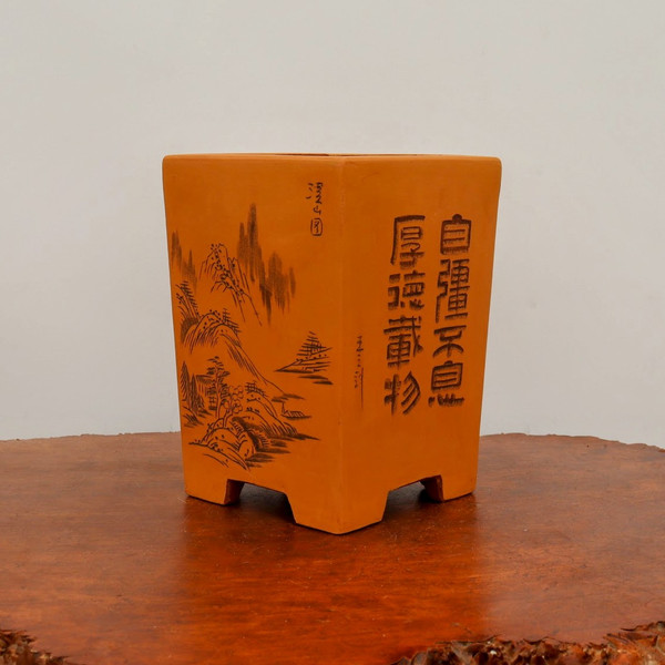 A square, unglazed terracotta bonsai pot sits on a polished burl wood base against a neutral white background. The pot features a tapered shape with four wide, blocky feet at the bottom. On the right side, two columns of large, etched Chinese characters stand out in dark ink. On the left side, a delicate landscape illustration is etched into the clay, showing traditional mountain peaks, tiered roofs of small houses, and a cluster of trees. Fine calligraphic signatures and seals are subtly carved into the center and upper portions of the pot’s side, showcasing the artisanal craftsmanship of this decorative bonsai vessel.