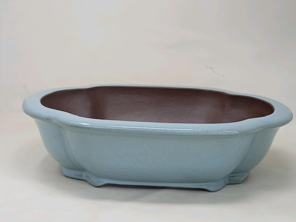 A side-angle view of an oval 16-inch Yixing bonsai pot, showcasing its elegant design and light blue glazed finish. The pot features a gently scalloped rim and a wide, shallow basin perfect for mature bonsai trees. While the exterior is coated in a smooth, powder blue glaze, the interior reveals a natural, dark brown unglazed clay characteristic of traditional Yixing pottery. The pot is supported by small, integrated feet that lift the base slightly off the ground, set against a clean, neutral background.