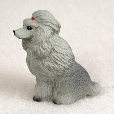 A detailed miniature figurine of a silver-gray poodle, designed as a decorative accent for a bonsai tree arrangement. The poodle is captured in a sitting profile, facing left, with its characteristic long, flowing hair on its head and chest sculpted in fine detail. A small, vibrant red bow sits atop its head, adding a pop of color to the monochromatic figure. The lower half of its body has a more textured, curly appearance. The figurine is placed on a light, sandy surface, illustrating how it might look as a charming addition to a miniature garden or bonsai landscape.