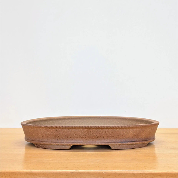 An oval, shallow, handmade bonsai pot crafted by artist Jack Hoover. The pot is made of an unglazed, reddish-brown clay with a natural, finely speckled texture. A distinctive rope-like decorative border runs along the outer edge of the upper rim. The body of the pot tapers slightly inward toward the base, which features a recessed panel and four integrated feet. These feet create small arched openings at the bottom for drainage and air circulation. The pot is centered on a light-colored wooden surface against a plain, off-white background, highlighting its earthy tones and artisanal craftsmanship.