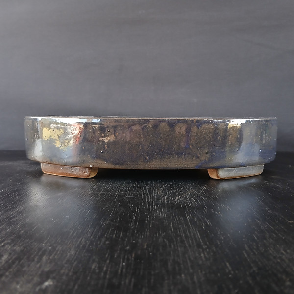 A profile view of a 12-inch glazed oval Yixing bonsai pot resting on a dark, wood-grain surface against a plain, dark gray background. The pot features a rich, dark glaze with a metallic, iridescent quality that reflects tones of deep blue, charcoal, and mottled patches of gold and bronze. Its low-profile, oval form has smooth, rounded edges and a level rim. The vessel is elevated on four small, unglazed rectangular feet in a light tan clay, which provides a natural contrast to the dark, glossy body of the ceramic. The lighting catches the reflective surface of the pot, highlighting its handcrafted texture and the depth of its glaze.