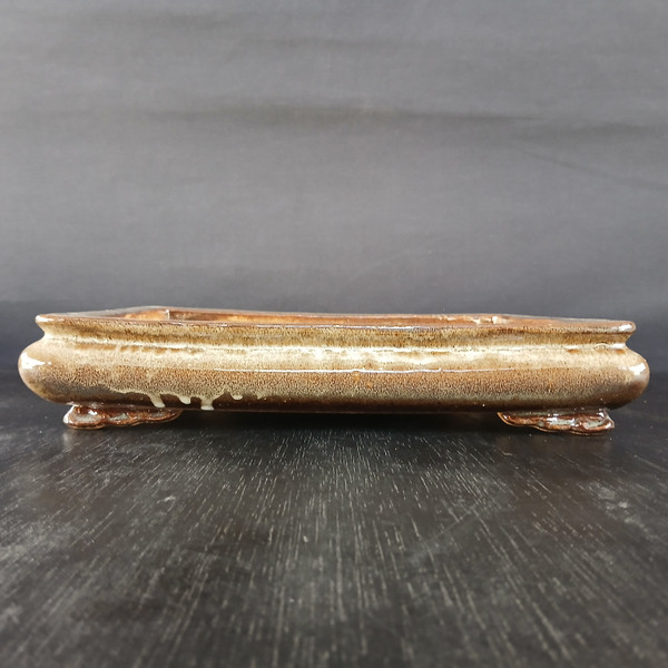 A side-view photograph of a 12-inch shallow rectangular Yixing ceramic bonsai pot (Model No. 2534f). The pot is finished with a glossy, variegated glaze in earthy shades of brown, tan, and cream, featuring a subtle mottled texture and a decorative drip of lighter glaze along the bottom edge. It has rounded corners and a low-profile design, supported by four small, subtly carved feet. The pot is positioned on a dark, wood-grained surface against a solid, dark gray background, emphasizing its traditional craftsmanship and elegant proportions.