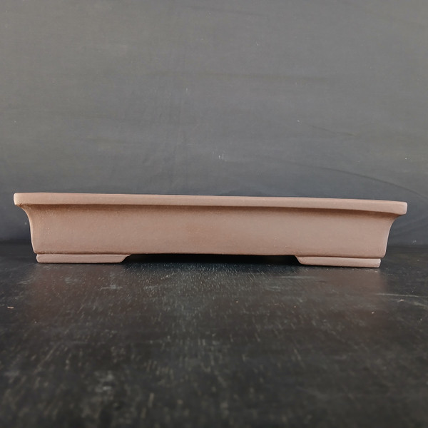 A side-profile photograph of a 14-inch unglazed rectangular Yixing bonsai pot. The pot is crafted from a smooth, matte, reddish-brown clay, characteristic of traditional Yixing pottery. It features a shallow design with a slightly flared upper rim and clean, straight lines that taper slightly toward the base. The pot is displayed on a dark, textured surface against a neutral dark gray backdrop, highlighting its minimalist and elegant architectural shape. The subtle recessed feet at the base provide a slight lift, making it a functional and aesthetic choice for showcasing mature bonsai trees.