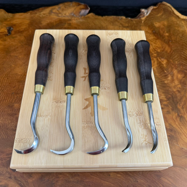 A professional 5-piece bonsai deadwood carving gouge set is displayed on its light-toned bamboo presentation case. The tools are arranged neatly in a row, showing a variety of curved and hooked blades designed for intricate wood carving. Each tool features a smooth, ergonomically shaped handle made of dark, polished wood, which is joined to the metal shank by a bright brass ferrule.

The metal shanks terminate in different gouge shapes—some with tight, narrow hooks and others with wider, more open curves—providing the versatility needed for creating jin and shari on bonsai trees. The bamboo presentation case has subtle Asian characters engraved into its surface and rests upon a large, polished slab of natural wood with a rich, swirling grain pattern in warm amber and brown tones. The composition is captured from a top-down angle, highlighting the craftsmanship and fine materials of both the carving tools and their storage box.