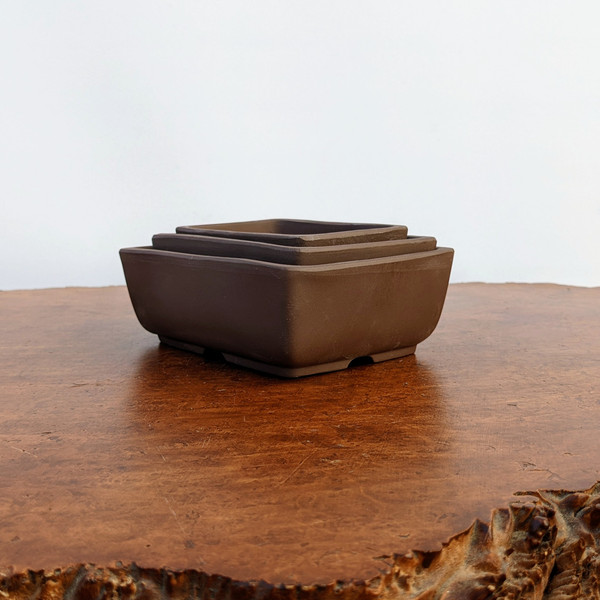 A nested set of three Udei brown unglazed ceramic bonsai pots, ranging from small to large, stacked inside one another. The pots feature a deep, earthy chocolate-brown color with a smooth, matte finish characteristic of unglazed clay. Each pot has a rectangular shape with slightly flared sides and subtly rounded corners. The largest pot at the bottom shows small drainage or ventilation cutouts at its base. The set is displayed on a polished, warm-toned natural wood slab with a visible grain and a rustic live edge in the foreground, all set against a clean, minimalist white background.