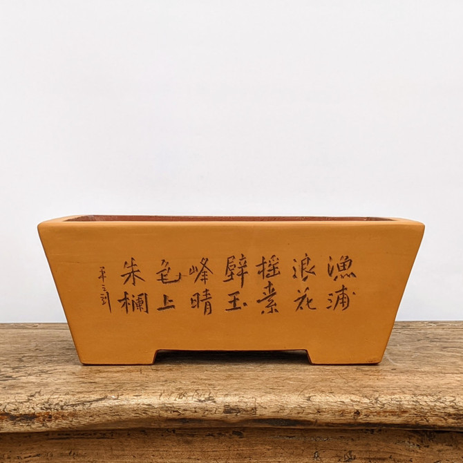 An eye-level, front-view photograph of a rectangular, unglazed ceramic bonsai pot made from warm, ochre-colored clay. The pot features a traditional tapered shape, narrowing slightly from the top rim down to a sturdy base with integrated feet visible as cutouts. The centerpiece of the design is a series of hand-etched Chinese calligraphy characters across the front face, rendered in a dark, contrasting tone that highlights the fine carving. The pot sits centrally on a rustic, weathered wooden surface with a prominent grain, all positioned against a stark, plain white background that emphasizes the earthy tones and textures of the unglazed stoneware.