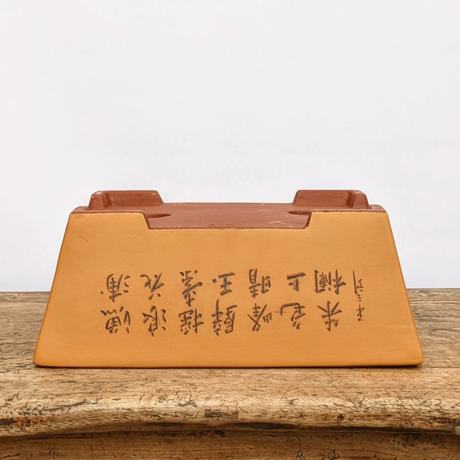 A detailed photograph of a rectangular unglazed bonsai pot, resting upside down on a rustic, weathered wooden surface against a solid white background. The pot features a two-tone color scheme: a smooth, matte ochre-yellow body with a deeper terracotta red on the top rim and feet. The focal point of the image is the hand-etched Chinese calligraphy, arranged in two rows of traditional characters across the side of the pot, with a smaller vertical signature to the right. The pot's bottom, facing upward, reveals circular drainage holes, highlighting its functional design for bonsai cultivation. This image showcases the artistic craftsmanship and traditional aesthetic of an etched ceramic bonsai container.