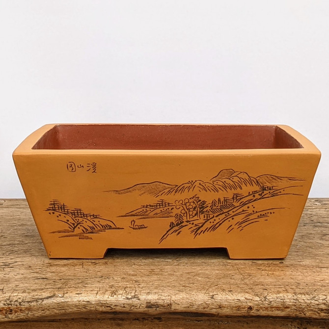 A rectangular, unglazed ceramic bonsai pot in a warm orange-brown terracotta color, resting on a rustic wooden table. The front face of the pot features a detailed, hand-etched landscape scene in a traditional East Asian style. The scene depicts serene rolling mountains in the distance, a small boat on a body of water, and a foreground filled with scattered trees, small huts, and winding paths. In the upper left corner of the scene, there are three small Chinese characters etched into the clay. The pot has a clean, flat top rim and slightly tapered sides that lead down to small, integrated feet. This classic bonsai container is designed to complement the natural beauty of a miniature tree with its earthy tones and intricate, hand-crafted artwork.