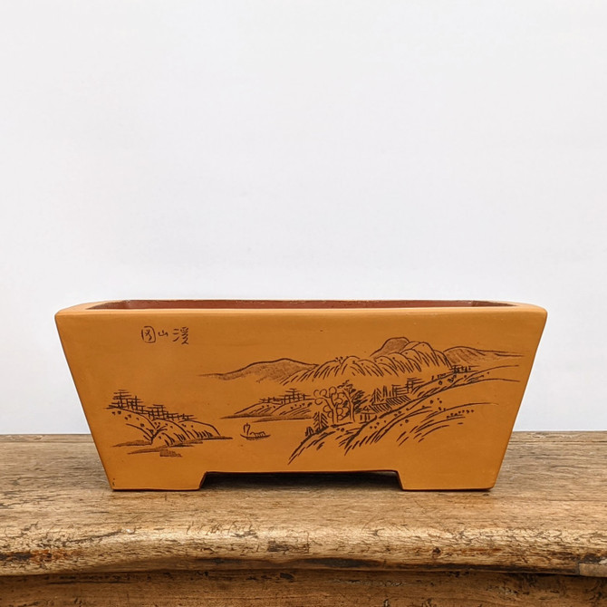 An unglazed rectangular bonsai pot made from warm, terracotta-colored clay sits prominently on a weathered wooden tabletop. The pot’s front face features a delicate, hand-etched landscape in a traditional East Asian style, depicting rolling hills, clusters of small buildings or huts, and a lone sailboat on a tranquil body of water. In the upper left corner of the scene, three Chinese calligraphy characters are neatly inscribed. The pot has a slightly flared rim and sturdy, blocky feet that elevate it from the rustic wood surface. The entire piece is set against a clean, minimalist off-white background, highlighting the fine craftsmanship and earthy texture of the ceramic.