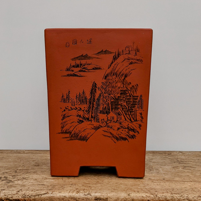 A tall, tapered rectangular unglazed bonsai pot, crafted from a warm, reddish-brown terracotta-style clay, stands on a weathered wooden surface. The front of the pot features a detailed, traditional black ink-style landscape etching. The scene depicts a serene mountain range with rolling hills, textured pine trees, and small cottages nestled into the slopes. Above the landscape in the upper left corner, four traditional Chinese characters are inscribed alongside a small, circular red seal mark. The pot has a deep, functional design ideal for cascade or semi-cascade bonsai styles, with two subtle feet carved into the base for drainage and stability. The earthy texture of the ceramic is highlighted against a clean, neutral gray background, emphasizing its rustic and classic aesthetic.