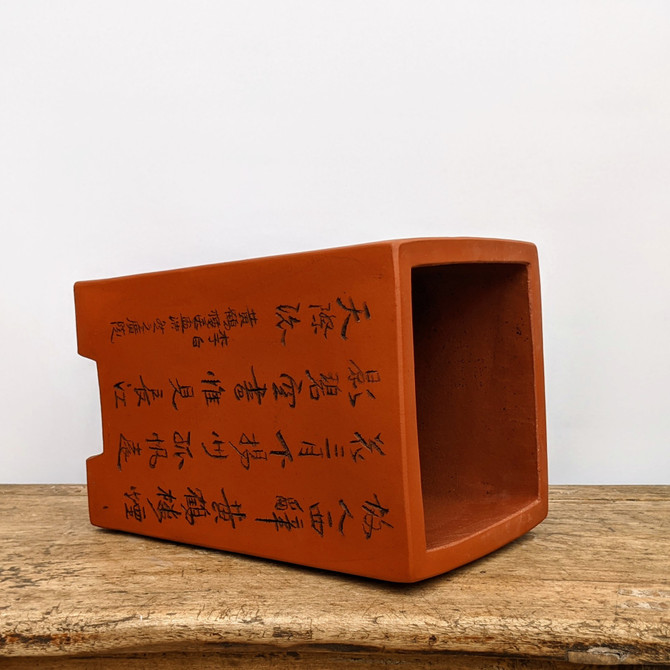 An unglazed, reddish-brown rectangular bonsai pot crafted from traditional clay, resting on its side on a rustic, weathered wooden plank. The side of the pot features intricate, hand-inscribed black Chinese calligraphy. The image highlights the pot’s smooth matte texture, its slightly tapered rectangular shape with small blocky corner feet, and its empty interior. Set against a clean, white background, this elegant ceramic container showcases a blend of functional bonsai design and classic Eastern artistry.