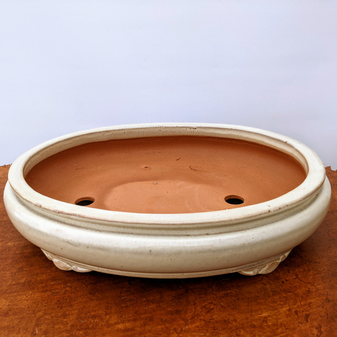 An eye-level, slightly high-angle studio shot features a shallow, oval glazed bonsai pot. The pot has a smooth, creamy white exterior glaze with a subtle glossy finish and fine, dark speckling. It sits on several small, decorative cloud-style feet. The interior of the pot is unglazed, showing the natural, porous, reddish-brown terracotta clay. Two large, circular drainage holes are visible at the bottom of the pot, essential for proper water drainage and root health in bonsai cultivation. The pot rests on a textured, warm-brown surface, possibly wood or cork, against a plain, off-white background that emphasizes the pot's clean lines and elegant design.