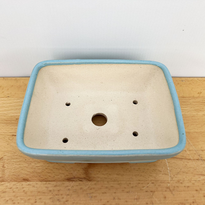 A top-down, angled view of a 7-inch rectangular bonsai pot featuring a light blue glaze on its rounded rim and exterior. The interior of the ceramic pot is unglazed, showing a natural matte tan color. For functionality, the bottom of the pot has a large circular drainage hole in the center and four smaller wiring holes, one positioned near each corner, to allow for secure plant anchoring. This Yixing bonsai pot is resting on a light-toned wooden surface against a plain, off-white background.