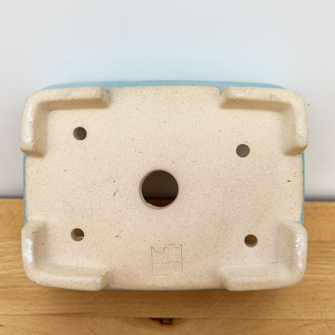 An overhead shot of the underside of a 7-inch light blue glazed Yixing bonsai pot, showcasing its functional design. The unglazed bottom is made of light-colored, speckled beige clay and features five circular drainage holes: one large hole positioned in the center and four smaller ones located near each corner to ensure proper water drainage. 

The pot is supported by four integrated, L-shaped raised feet at the corners, providing necessary elevation for airflow. A square artist's chop or seal is clearly impressed into the clay near the bottom edge, indicating its provenance. A thin rim of the smooth, light blue glaze is visible along the outer edges of the rectangular pot. The item is resting on a light-grained wooden surface against a plain, off-white wall.