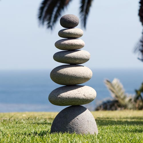 A tall, balanced rock cairn featuring seven smooth, naturally rounded stones stacked vertically from largest to smallest. The stones range in color from cool grey to warm beige, showcasing a natural weathered texture and soft shadows from the sunlight. The stack begins with a substantial, dome-shaped base resting on lush green grass and culminates in a small, spherical top stone. The background features a serene, out-of-focus coastal scene with a blue ocean horizon and soft palm fronds, highlighting the cairn's role as a Zen-inspired garden accent or meditative outdoor focal point.