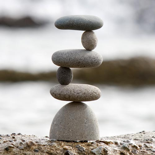 A photograph of an artistic side-to-side rock cairn, showcasing six smooth, grey river stones expertly balanced in a tall, precarious zigzag formation. 

The sculpture begins with a large, rounded stone at the bottom, resting on a rough, textured ledge. Balanced on top of it is a flat, horizontal oval stone, followed by a smaller, dark, egg-shaped stone standing vertically on its end. The stack continues with another flat stone shifted to the right, a small round stone standing on the right edge, and finally, a long, flat stone balancing horizontally at the very top. 

The alternating arrangement of horizontal and vertical stones creates a striking "Z" shape, emphasizing a sense of zen-like balance, stability, and tranquility. The background is a soft-focus, blurred vista of a tranquil beach or shoreline, allowing the intricate detail and impressive composition of the stacked rocks to stand out as a piece of natural garden decor.
