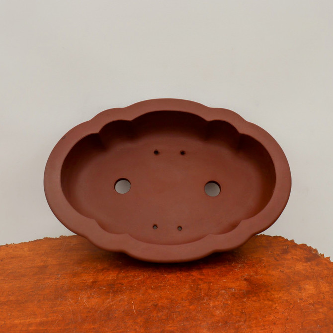 A top-down view of a professional-grade, unglazed Yixing bonsai pot in a deep reddish-brown clay. The pot features a distinctive scalloped or lobed oval rim, creating an elegant cloud-like silhouette. Inside the smooth, matte interior, two large circular drainage holes and four smaller wire holes are visible at the bottom, essential for healthy tree growth and stability. The pot is displayed on a textured, warm-toned wooden surface against a neutral, off-white background, emphasizing its classic aesthetic and functional design for bonsai enthusiasts.