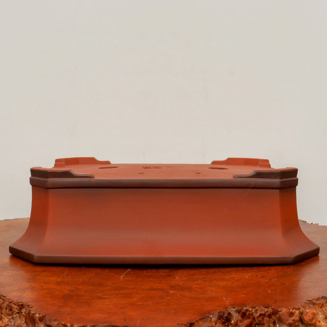 An eye-level, close-up shot of an unglazed, reddish-brown yixing clay bonsai pot, shown upside down to highlight its construction and drainage features. The pot has a rectangular shape with flared sides and clipped corners that form an octagonal footprint. The bottom surface, facing upwards, displays several small drainage holes and a subtle rectangular maker's mark stamped in the center. The clay has a smooth, matte texture with slightly darker highlights along the edges and corners. The pot is resting on a highly polished, warm-toned wood stand with a rustic, natural edge, all set against a clean, off-white background. This image showcases the craftsmanship and functional details of a classic unglazed bonsai container.