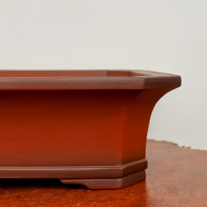 A detailed side-profile view of a rectangular unglazed bonsai pot with clipped or octagonal corners. The pot is crafted from a smooth, finely textured reddish-brown clay, showcasing a warm, earthy color. It features a wide, flat rim that flares outward, transitioning into sides that gracefully curve inward toward the base. The bottom is accented with a decorative tiered or stepped design and small integral feet. The pot is shown resting on a dark, textured wood surface against a plain, neutral off-white background, emphasizing its clean, elegant lines and the artisanal craftsmanship of the unglazed ceramic.