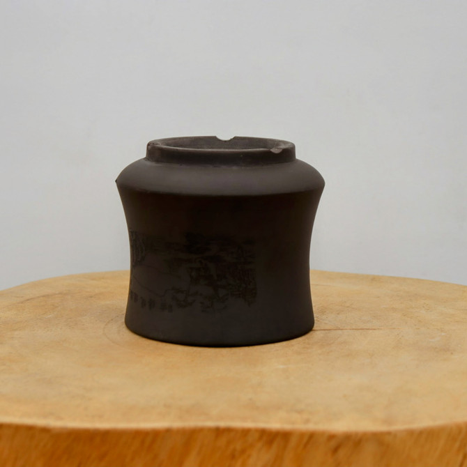 An unglazed, charcoal grey ceramic bonsai pot sits atop a natural wood pedestal. This small, artisanal pot features a rounded, slightly tapered body with a prominent shoulder and a narrow, notched rim. On its side, a detailed landscape scene is delicately etched, depicting mountain silhouettes and traditional scenery, with subtle calligraphic characters visible near the base. The pot rests on a thick, circular cross-section of a light-colored wooden log that displays its natural grain and a single radial crack. The entire composition is set against a clean, off-white background, emphasizing the pot's dark color and the fine craftsmanship of its etched design.