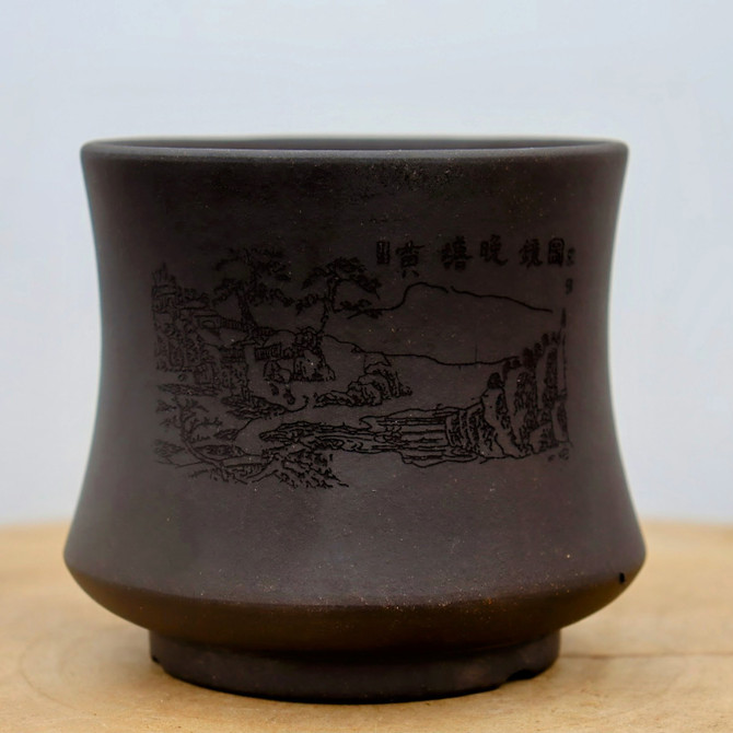 A close-up of a small, deep unglazed bonsai pot crafted from dark charcoal-brown clay with a matte finish. The pot features a unique shape that tapers upward from a sturdy base before flaring out slightly at the top rim. 

The front of the pot is decorated with a detailed, fine-line etching of a traditional East Asian landscape, depicting gnarled pine trees, rolling hills, and a serene valley. To the upper right of the mountain scene, four Chinese calligraphy characters are etched into the clay. The pot sits on a light-toned wooden surface against a neutral, off-white background, highlighting its earthy texture and artisanal design.