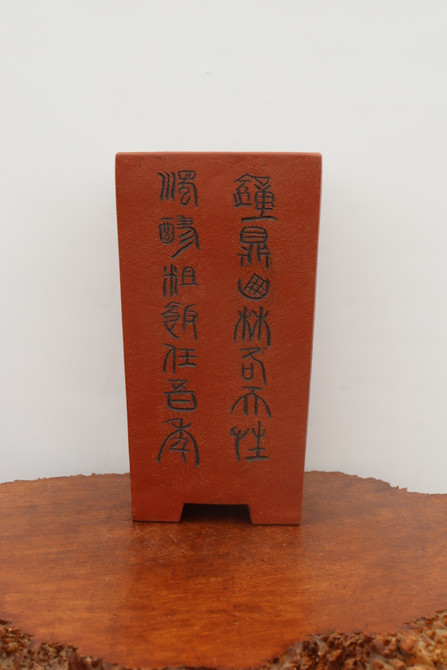 An eye-level, front-facing studio shot of an elegant, tall rectangular unglazed bonsai pot. The pot is crafted from a rich, reddish-brown clay with a fine, matte texture. The centerpiece of the design is two vertical rows of dark-etched Chinese calligraphy characters running down the front face. The pot tapers slightly towards the base and stands on two small block feet, creating a subtle arch underneath. It is displayed on a rustic, polished wood slab with visible grain, set against a plain, minimalist white background to highlight the pot's artisanal details and traditional aesthetic.