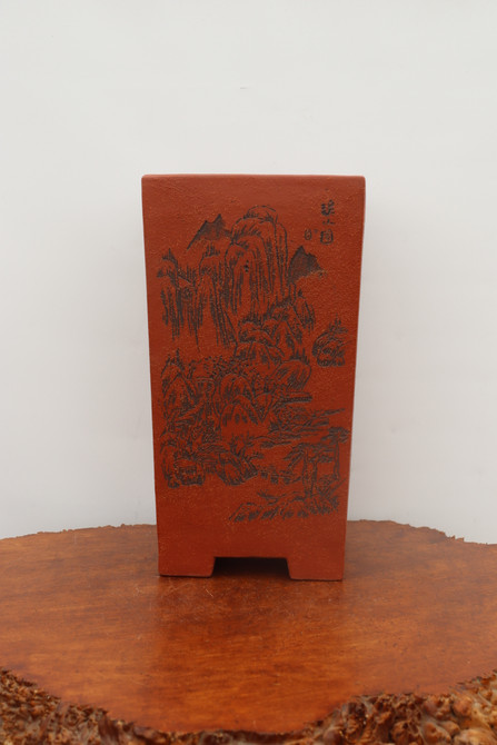 A tall, rectangular unglazed bonsai pot made from a textured, reddish-brown clay, ideal for cascade-style bonsai. The front face of the pot features an intricate, hand-etched landscape in black, depicting traditional mountain peaks and gnarled pine trees in an East Asian ink-wash style. Small Chinese calligraphic characters are etched in the upper right corner of the art. The pot has a slightly tapered profile and sits on four low, integrated feet. It is displayed on a polished, warm-toned natural-edge wooden burl slab against a clean, off-white wall, highlighting its artisanal quality and traditional aesthetic.