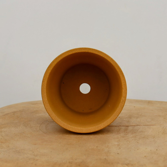 A top-down view of a small, round unglazed ceramic bonsai pot in a warm ochre or light orange color. The pot features a thick, smooth rim and a single circular drainage hole at the center of its base. It is resting on a light brown, rustic wooden surface with a visible natural grain, set against a plain, neutral-toned off-white background.
