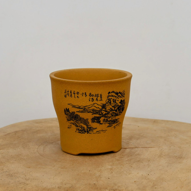A high-angle, eye-level shot features a small, unglazed bonsai pot with a warm, yellow-ochre finish. The ceramic pot has a cylindrical body that tapers slightly toward the base, featuring a subtly flared rim and four small, integrated feet. Centered on its front is a delicate black-ink etched landscape, depicting a tranquil scene of rolling mountains, clusters of pine trees, and a small traditional pavilion nestled near a body of water. To the left of this scene, several vertical columns of elegant Chinese calligraphy are finely inscribed. 

The pot's matte, earthy texture and rich golden-brown color are highlighted by soft, even lighting that casts minimal shadows. It sits on a rustic, light-toned wooden surface with visible grain and natural cracks, adding to the organic feel of the composition. The entire setup is placed against a clean, neutral off-white background, creating a minimalist aesthetic that focuses attention on the detailed craftsmanship of the etched artwork.
