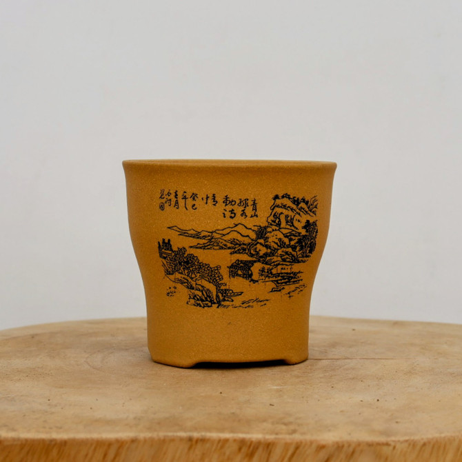 An ochre-colored, unglazed ceramic bonsai pot sits atop a natural wooden platform against a plain, off-white background. The pot features a classic, slightly tapered shape with a subtle flare at the rim and small, integrated feet at the base for drainage and airflow. Its matte, earth-toned surface is decorated with a detailed black etched or printed traditional Chinese landscape. The artwork depicts a serene scene with towering, craggy mountains, a calm lake, and small village huts nestled among clusters of trees. Above the landscape, a line of elegant Chinese calligraphic characters is inscribed horizontally, adding a touch of classic sophistication. The overall design emphasizes a minimalist yet artistic aesthetic, characteristic of high-quality unglazed bonsai containers.