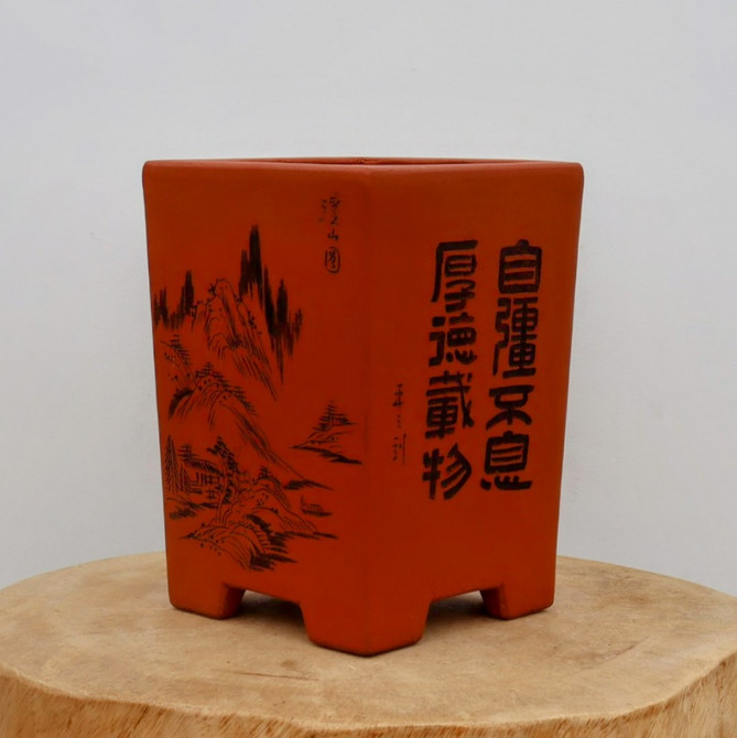 A tall, square, unglazed bonsai pot crafted from red terracotta clay, featuring intricate black etched designs on its exterior facets. On the left side, a traditional landscape scene is depicted with fine lines showing jagged mountain peaks, scattered pine trees, and small dwellings nestled into the hills. The right side displays bold, vertical Chinese calligraphy characters etched in black. The pot has a matte, earthy texture and is supported by four short, blocky feet. It is displayed on a circular, light-colored wooden slab against a plain white background, highlighting the contrast between the deep red of the clay and the black etched artwork.