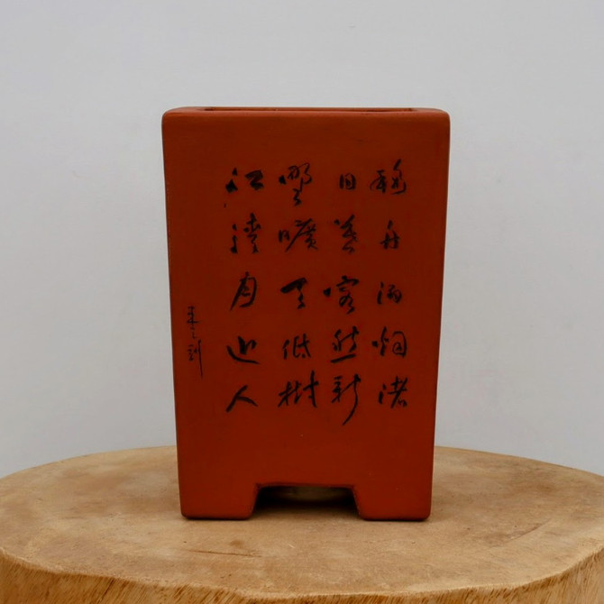 An upright, rectangular unglazed bonsai pot made of rich, reddish-brown terracotta clay. The pot features a tall, slightly tapered silhouette with two rectangular feet at the base. Its front face is decorated with four vertical columns of hand-etched Chinese calligraphy in black ink, accompanied by a smaller vertical signature on the left side. The pot has a smooth, matte finish characteristic of high-quality unglazed stoneware. It is displayed on a rustic, circular cross-section of a light-colored wooden log against a neutral, off-white background, highlighting the traditional craftsmanship and earthy aesthetic of the artisan-etched container.