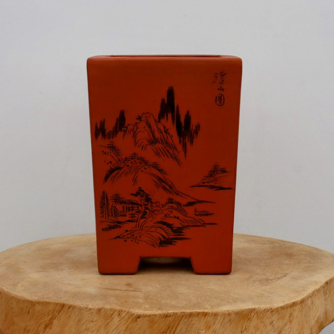 An eye-level, front-facing shot captures a rectangular, unglazed ceramic bonsai pot in a warm, reddish-brown terracotta color. The pot has a deep, square profile that tapers slightly toward the base and is supported by four integrated block-style feet. Its front surface features a delicate, black-ink etched landscape in a traditional East Asian style, depicting towering, craggy mountain peaks, scattered pine trees, and a small rustic hut nestled at the base of the hills. Small black calligraphic characters are visible in the upper right corner of the etched scene. The bonsai pot sits centered on a thick, natural wood slice base with a light-colored grain and rough bark edges, all set against a clean, neutral-white background that highlights the pot's earthy tones and detailed artwork.