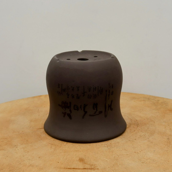 A studio-lit, eye-level shot captures an upside-down unglazed bonsai pot crafted from deep chocolate-brown clay. Resting on a light-colored, circular wooden pedestal, the pot’s flared, bell-shaped form is elegantly displayed. The base, which is positioned at the top in this view, features a large central drainage hole and several smaller wire holes for securing a tree, along with three subtle, built-in feet along its rim.

The middle section of the pot is intricately decorated with hand-etched Chinese calligraphy. The script varies in style, featuring large, expressive, flowing characters alongside rows of smaller, more precisely carved text. The matte finish of the unglazed ceramic highlights the fine details of the etchings and the smooth, earthy texture of the clay. The neutral off-white background draws focus to the pot’s traditional craftsmanship and sophisticated design.
