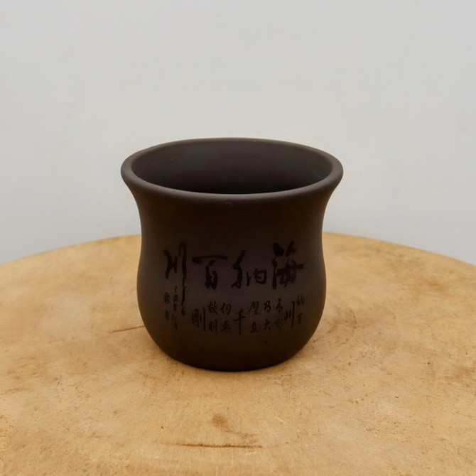A small, unglazed bonsai pot made of dark brown ceramic sits centered on a light-toned wooden surface. The pot has a round, bell-like shape with a gently flared rim and a smooth, matte finish. Its most striking feature is the intricate, traditional Chinese calligraphy etched into its side. The large, bold characters are accompanied by smaller lines of script arranged vertically, giving the piece a classic and artisanal feel. The pot is positioned against a clean, neutral-colored wall, while the wooden base it rests on displays natural grain patterns and a single, fine crack, adding a rustic element to the composition. The soft, even lighting emphasizes the pot's dark, earthy tones and the delicate details of the etched inscriptions.