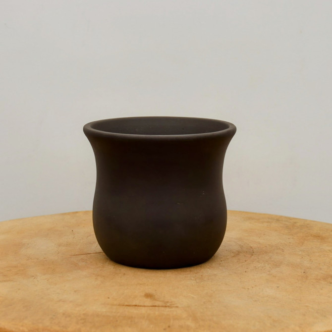 A small, dark brown unglazed ceramic bonsai pot sits centered on a light-toned wooden surface. The pot features a classic, elegant silhouette with a subtly flared rim that tapers inward at the neck and then curves outward into a rounded, bulbous lower section. Its smooth, matte finish is a deep chocolate brown, characteristic of traditional unglazed clay vessels used for bonsai. The pot is positioned on a circular, light-tan wooden slab, possibly a cross-section of a tree trunk, showcasing its natural grain. The entire scene is set against a plain, off-white background, creating a minimalist and clean aesthetic that emphasizes the pot's graceful form and earthy texture.