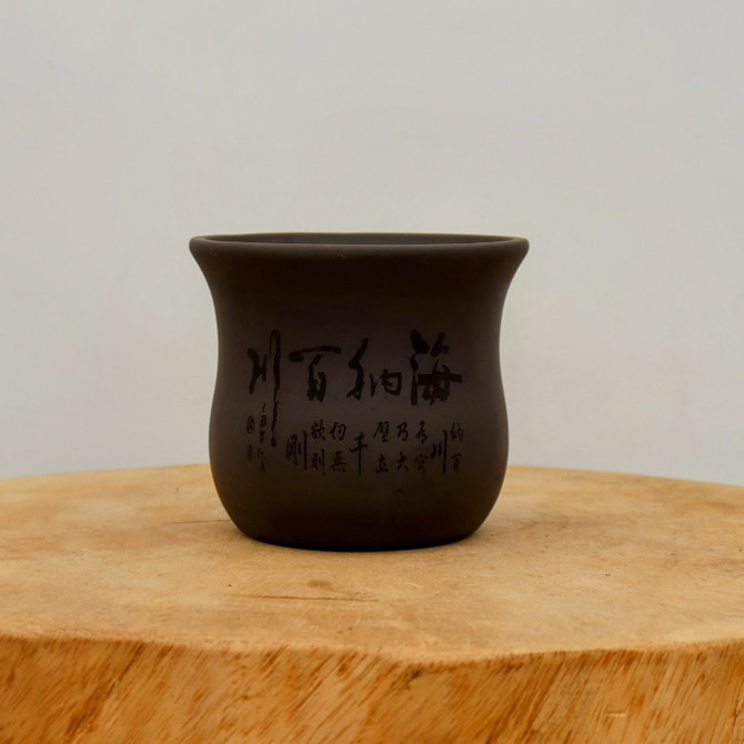 A studio-shot, eye-level close-up of a small, dark brown, unglazed ceramic bonsai pot. The pot features a classic design with a flared rim and a rounded body that tapers slightly toward the base. Its matte surface is adorned with intricate black Chinese calligraphy characters of varying sizes, which are etched into the side of the pot. A set of larger, bold characters dominates the center, while smaller, more delicate inscriptions and a traditional-style signature seal are positioned below and to the side. The pot sits on a thick, circular piece of light-toned natural wood, showcasing its organic grain and texture. The entire composition is set against a clean, neutral, off-white background, emphasizing the pot's earthy tones and artistic, etched details.