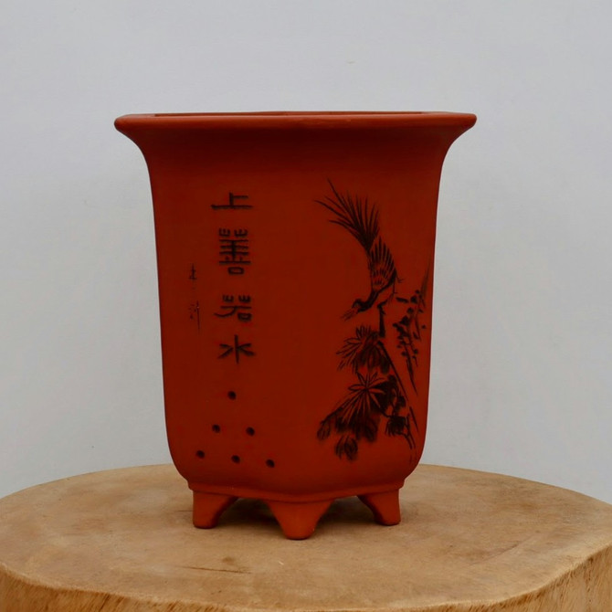 An eye-level, front-facing photograph shows a tall, rectangular unglazed terracotta bonsai pot resting on a thick, light-colored natural wood slab. The pot is a deep, earthy reddish-brown color with a flared top rim and four short, tapered feet at the base. 

The front surface of the pot is adorned with traditional black etched designs. On the right, a detailed illustration depicts a bird with long tail feathers perched atop a pine-like branch. To the left of the bird, four large Chinese characters are vertically inscribed: "上善若水" (Shàng shàn ruò shuǐ), a philosophical phrase meaning "The highest excellence is like water." A smaller vertical inscription, likely a maker's mark or signature, is visible further to the left. 

Near the bottom of the pot, a series of small, circular drainage and aeration holes are arranged in a triangular pattern. The entire scene is set against a clean, neutral off-white background, highlighting the pot's texture and artistic detail.