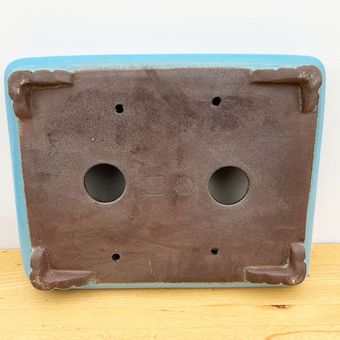 A bottom-up view of a 12-inch shallow rectangle Yixing ceramic bonsai pot, highlighting its functional design and authentic craftsmanship. The exterior of the pot is finished in a smooth, light-blue glaze, while the underside reveals the unglazed, dark-brown texture of the genuine Yixing clay. The base features two large, circular drainage holes centered for proper water flow, flanked by four smaller holes positioned near the corners for wiring a bonsai tree securely in place. Four sturdy, L-shaped feet provide elevation and stability at each corner. Two distinct impressed seals, one square and one circular, are visible in the center, representing the potter’s official marks of quality. The pot is resting on a light-toned wooden surface against a plain white background.